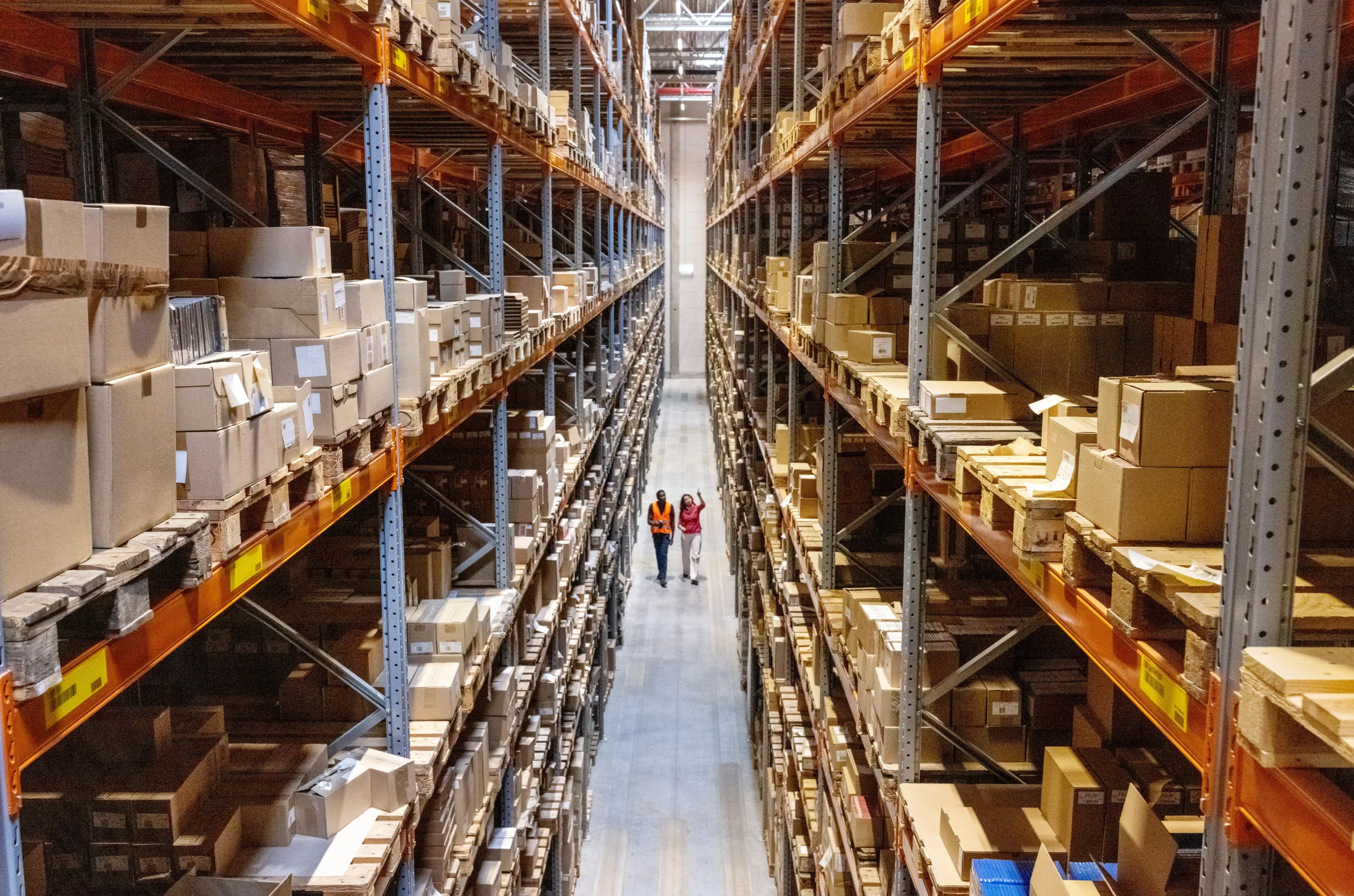 Workers walking through a large, organized warehouse aisle with tall shelving units filled with boxed inventory on both sides.